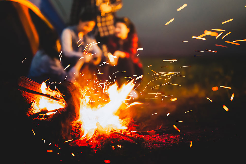 Im Vordergrund ein Lagerfeuer, im Hintergrund sitzen junge Leute und reden oder diskutieren